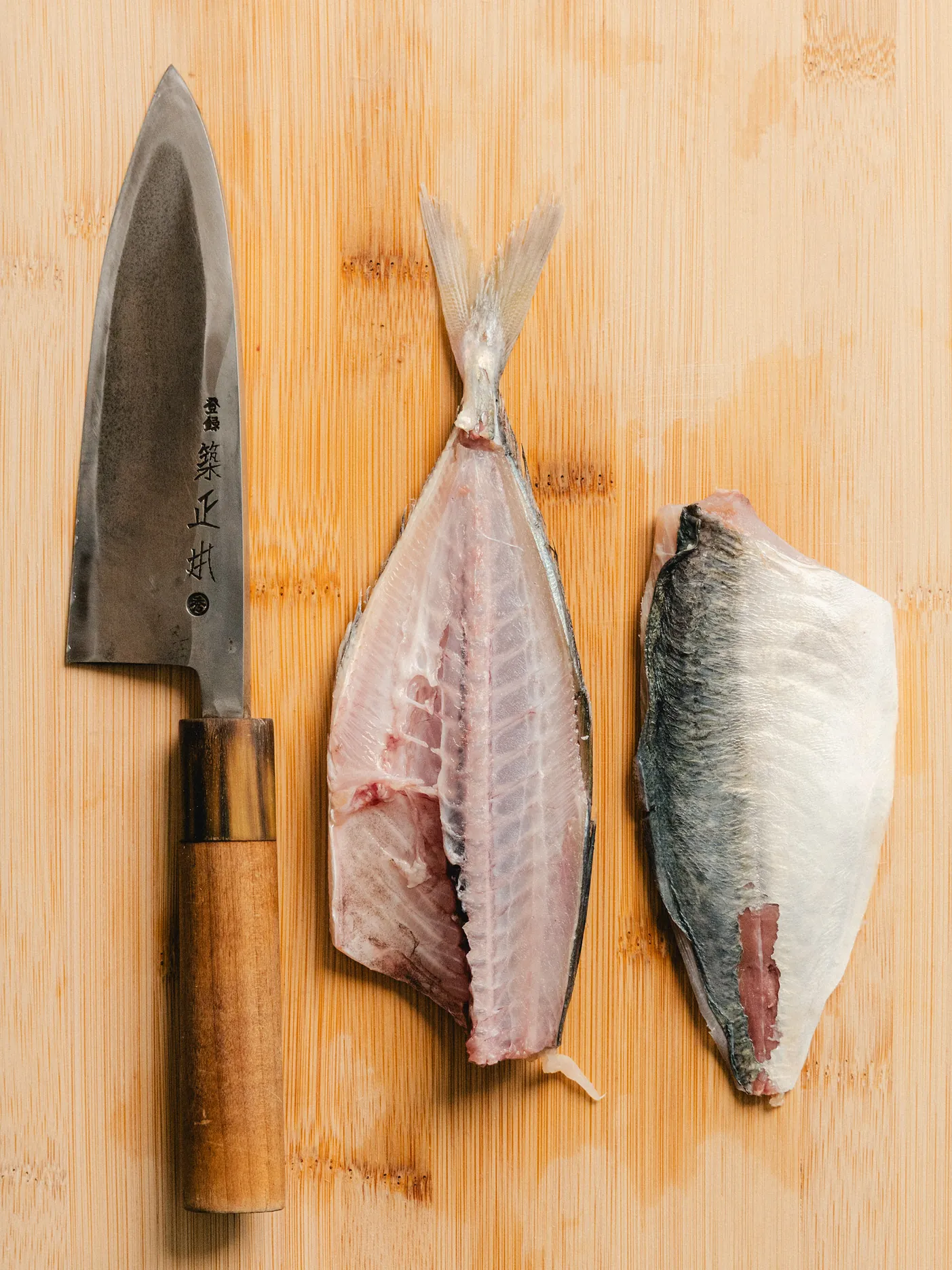 A Japanese knife beside two mackerel fillets on a bamboo board